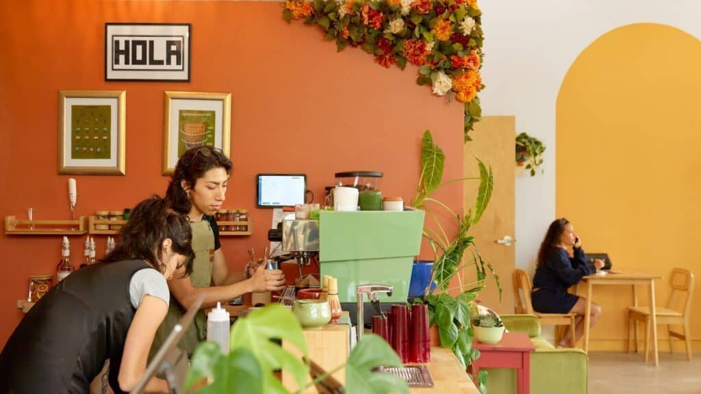 Two baristas work behind a counter in a brightly decorated café with orange walls, green plants, and a “HOLA” sign, while a woman sits alone at a table in the background.