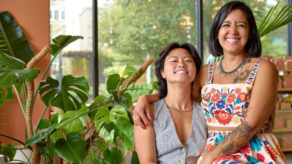 Two women sit close together and smile, surrounded by green plants and large windows in a bright indoor space.