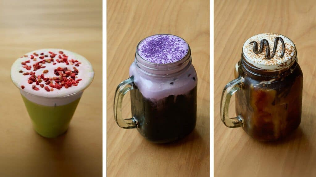 Three colorful specialty drinks in clear cups at Café Calaveras in the Central District: a green drink with dried berries, a purple drink with foam, and a dark drink topped with whipped cream and drizzle—perfect for celebrating our café opening.