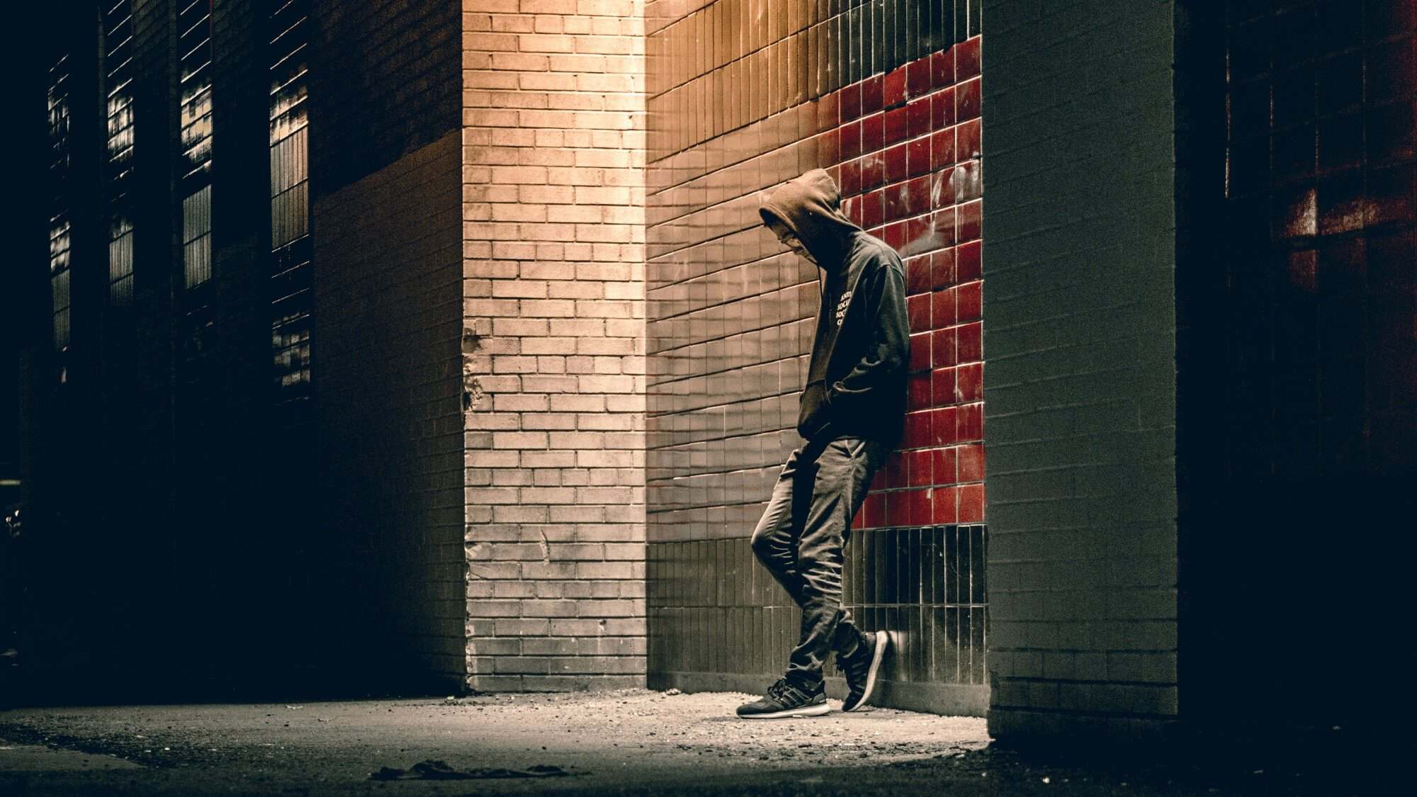 A person in a hoodie stands alone at night, leaning against a brick wall with red tiles, partially illuminated by a streetlight, capturing the quiet grief and boys' pain in the stillness.
