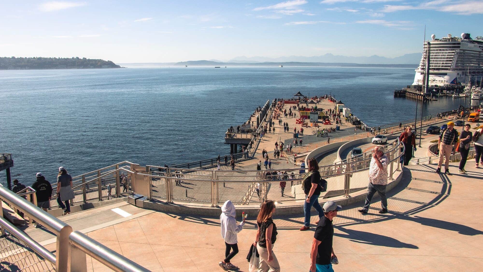 People walk along a waterfront promenade overlooking a pier extending into the water, with a cruise ship docked nearby under a clear sky.