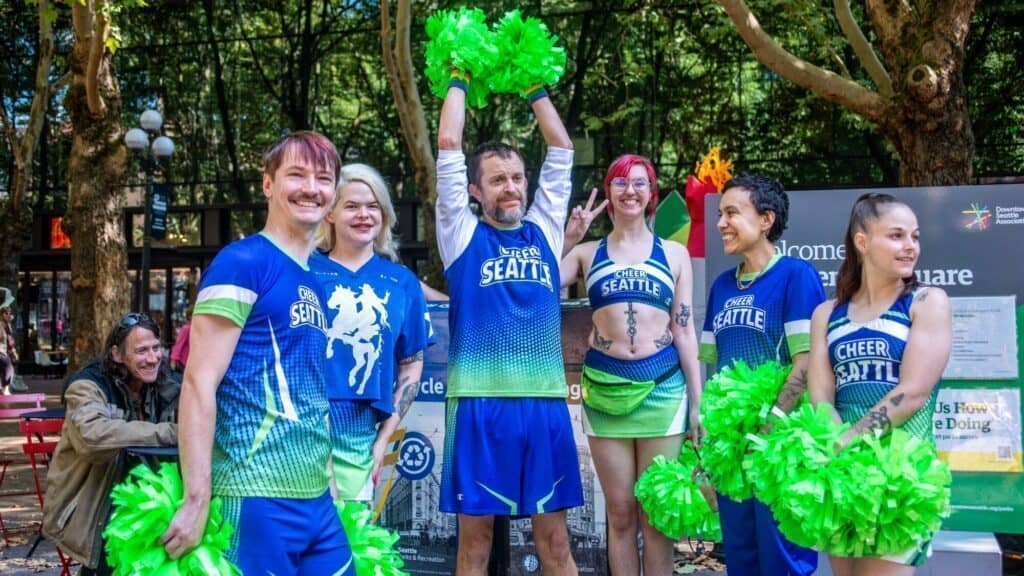 A group of six people in blue and green "Cheer Seattle" uniforms pose with green pom-poms outdoors in a park, with trees and informational signs in the background.
