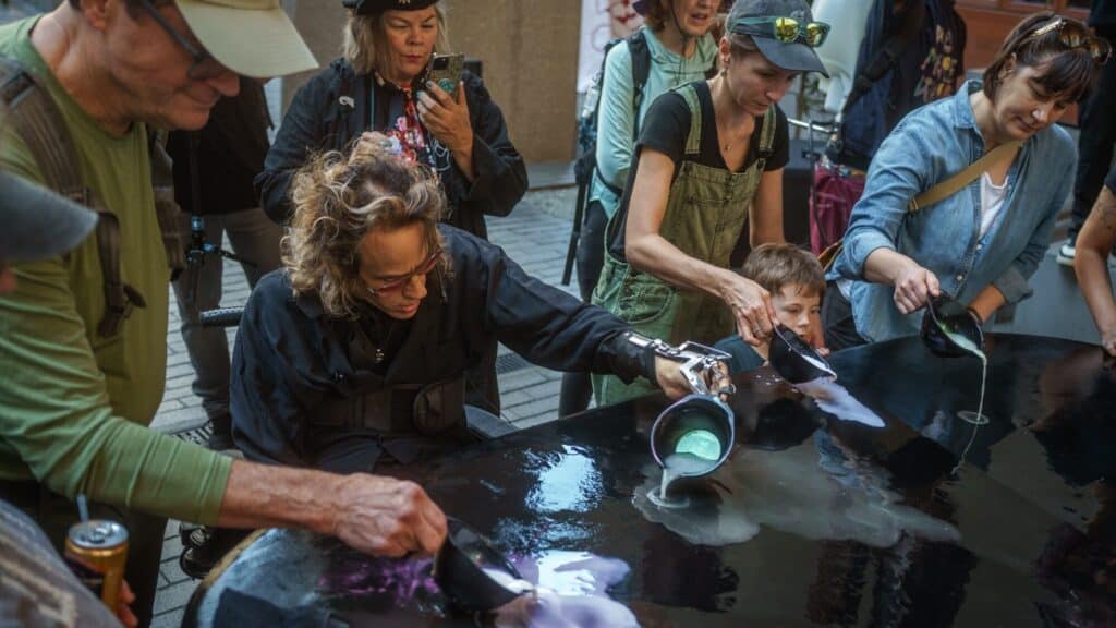 A group of people, including a child, pour colorful liquid from pitchers onto a reflective black surface at an outdoor event.