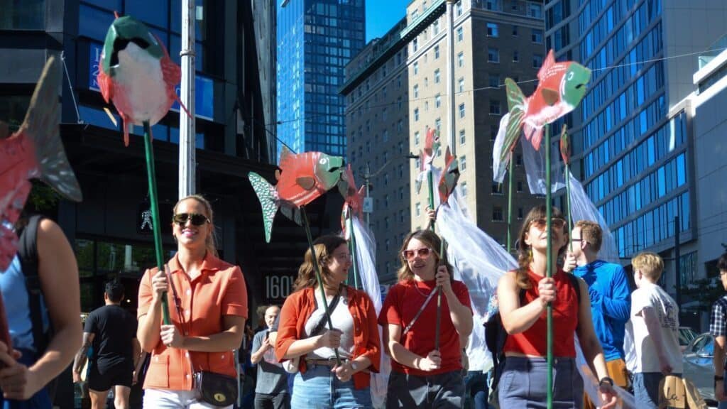 A group of people in casual clothing walk in Downtown Seattle, holding colorful fish-shaped puppets on poles during the sunny Seattle WALK DON'T RUN Festival.