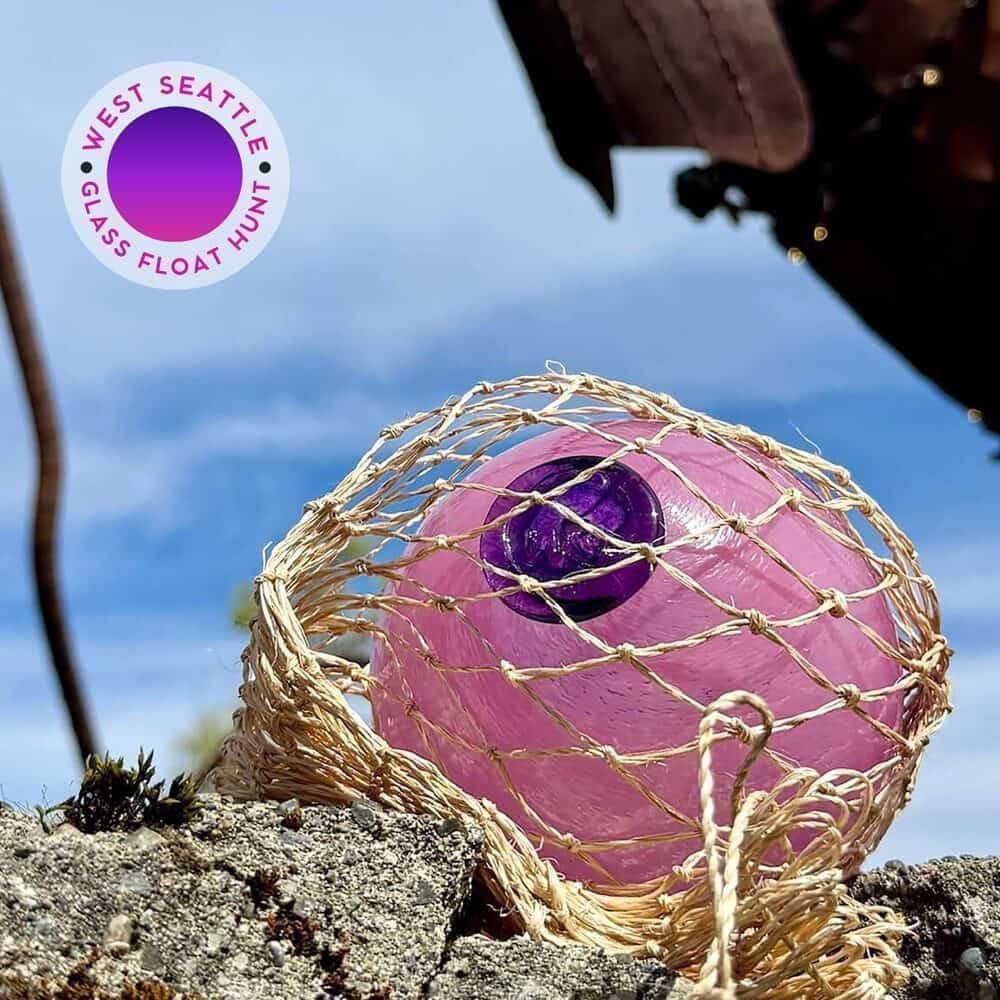 A purple glass float wrapped in netting rests on a rock outdoors, with a blue sky in the background. A circular "West Seattle Glass Float Hunt" logo is in the top left corner—add this to your Must List for 08-07-2025!.