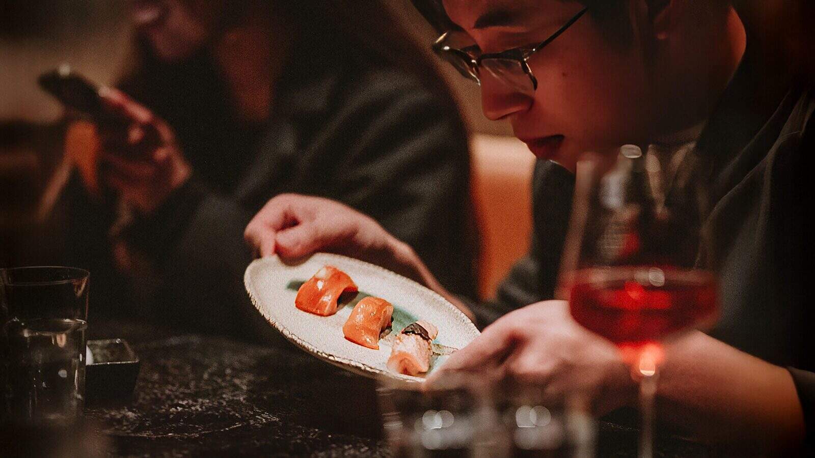 A person examines a plate of lab-grown salmon sushi at a dimly lit restaurant, with a wine glass and another person using a phone in the background.