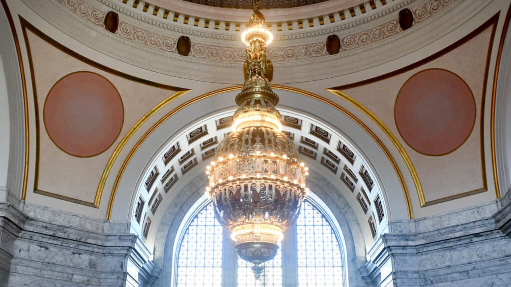 A large ornate chandelier hangs from the domed ceiling of a grand building with arched windows and decorative architectural details.